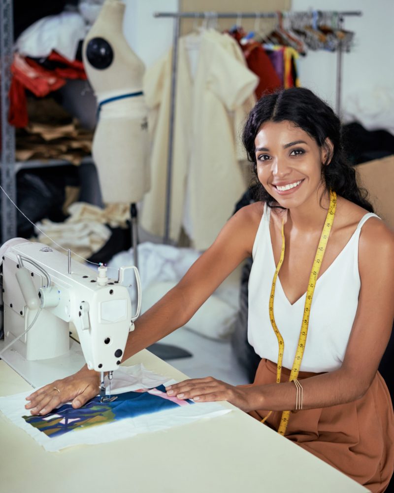 Portrait of seamstress sitting and smiling while sewing with professional machine at workshop